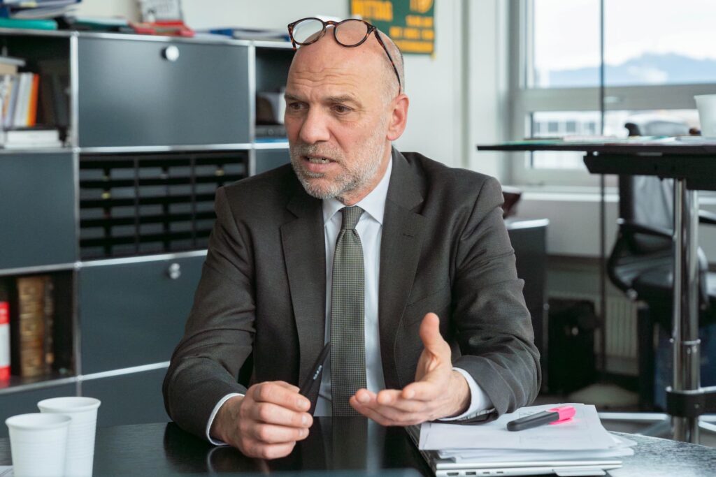 Florian Steinbacher, IV-Chef beim Bundesamt für Sozialversicherung im Gespräch am Tisch. Seine Hände gestikulieren. Er trägt einen dunklen Anzug und eine graue Kravatte. Er hat eine halbe Glatze, graues, sehr kurzes Haar und einen Bart.
Im Hintergrund ist das Büro zu sehen. Unter seinem Arm liegt ein Stapel Papier, auf welchem ein pinker Leuchtstift liegt.