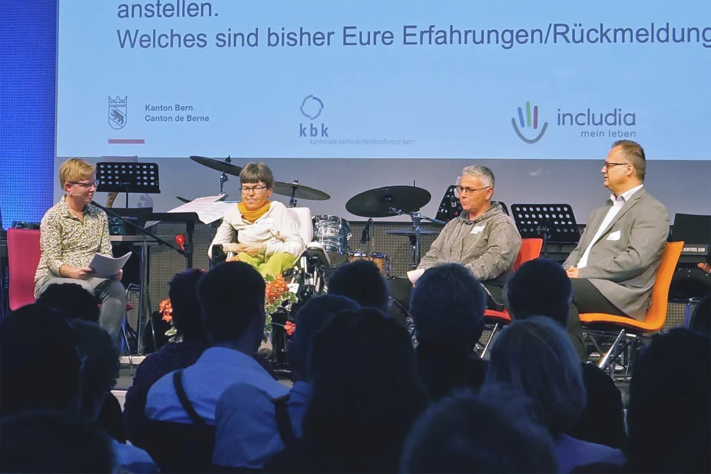 Ein Podium mit 4 Personen. Eine davon sitzt im Rollstuhl. Im Vordergrund sieht man verschwommen Zuschauende. Im Hintergrund leuchtet eine Präsentation, wo die 3 Logos vom Kanton Bern, der kantonalen Behindertenkonferenz kbk und von includia gut sichtbar sind. Alle wirken konzentriert.