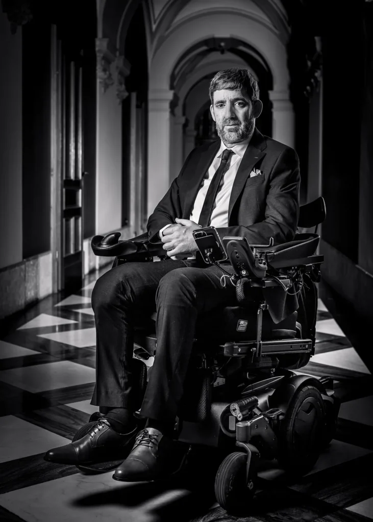 Eine Aufnahme in schwarz-weiss von Philipp Kutter im Rollstuhl in der Wandelhalle. Er trägt einen dunklen Anzug, dazu ein helles Hemd und eine dunkle Kravatte. Sein Blick ist ernst, wirkt eindringlich und geht direkt zur Kamera.
