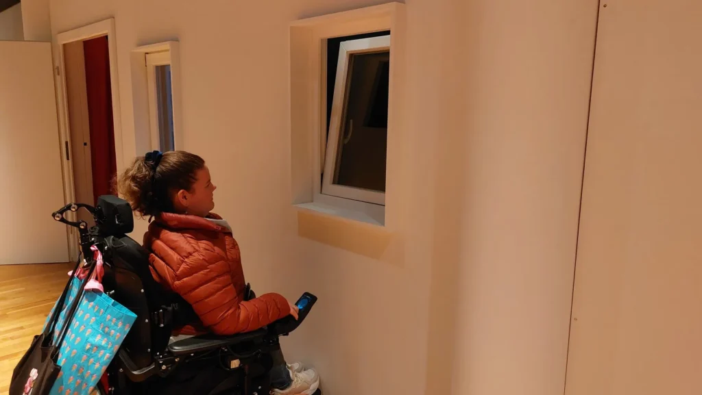 Eine Rollstuhlfahrerin steht vor einem Wand mit einem gekippten Fenster. Es handelt sich um eine Installation eines Museums, in welcher man durch das gekippte Fenster blicken muss. Die Person kann aber nicht nah und hoch genug an das Fenster heranfahren.
