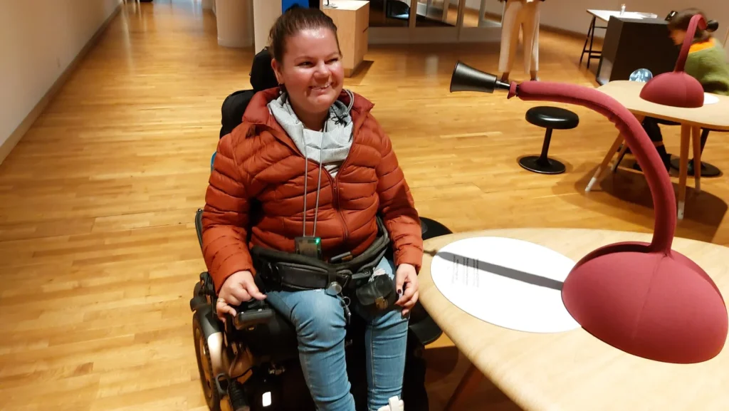 Eine Person sitzt in einem Rollstuhl. Sie trägt eine orangefarbene Daunenjacke und ein helles Halstuch. Sie lächelt. Sie steht/sitzt mit dem Rollstuhl vor einem Tisch. Dort befindet sich eine Installation, für welche man nah genug an den Tisch heranrücken können muss. Das ist im Rollstuhl nicht möglich.