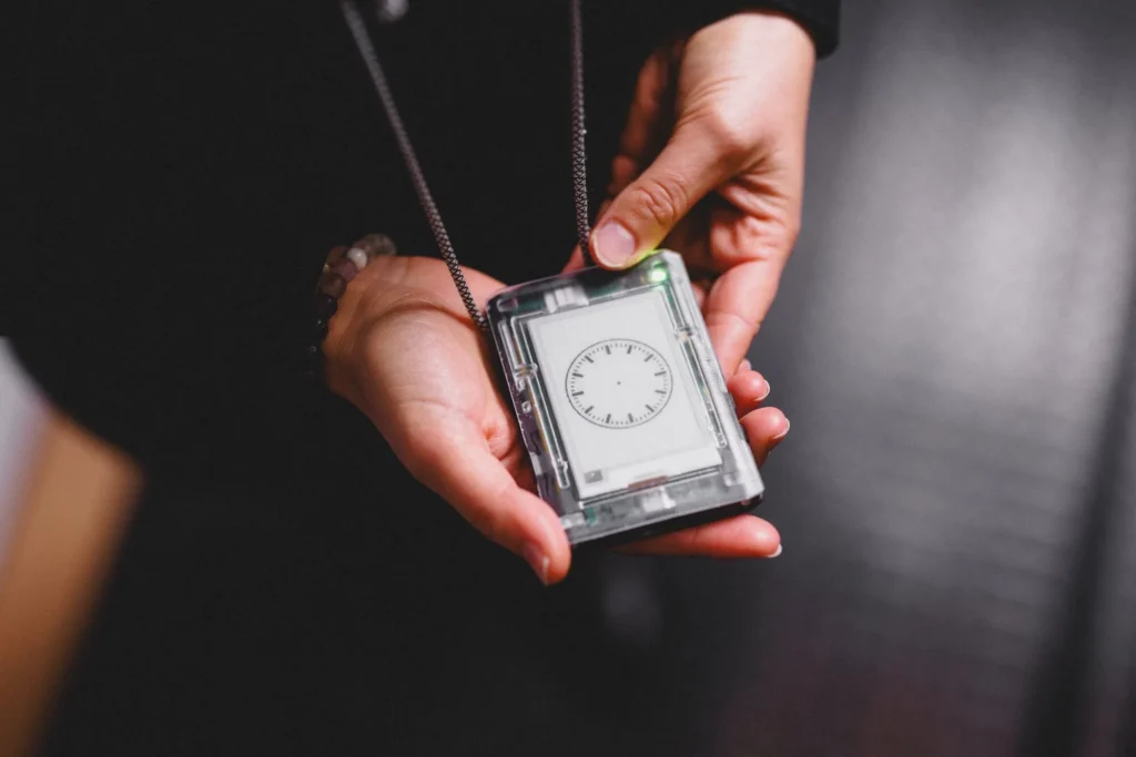 Zwei Hände, welche ein viereckiges Gerät - einen digitalen Guide - in den händen hält. Zwei Kordeln führen vom Gerät weg, vermutlich zum Hals der Person, die ihn hält. Auf dem digitalen Guide ist eine Art Uhr ohne Zeiger sichtbar. Ein Lämpchen leuchtet grün.